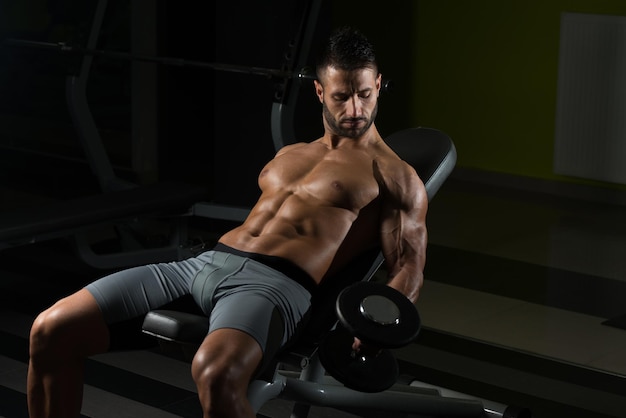 Bel homme travaillant sur les biceps dans un gymnase sombre boucles de concentration d'haltères