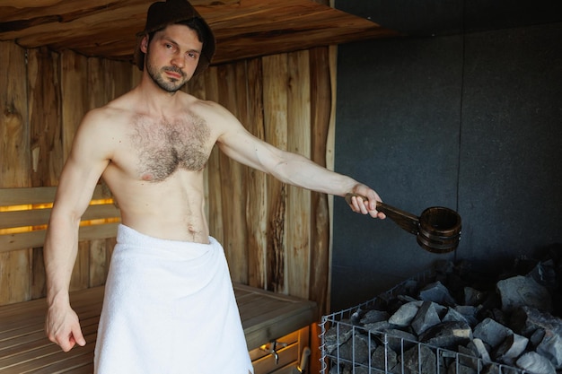 Bel homme versant de l'eau sur des pierres chaudes dans le sauna
