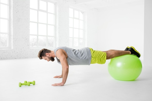 Bel homme en vêtements de sport faisant des pompes avec fitball à l'intérieur de la salle de sport blanche