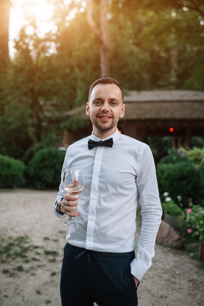 Un bel homme vêtu d’une chemise blanche et d’une cravate goûte le vin blanc dans un verre transparent.