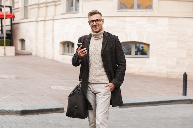 Bel homme vêtu d'un manteau marchant à l'extérieur, tenant un téléphone mobile