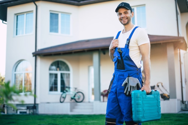 Un bel ouvrier confiant en uniforme spécial avec une boîte à outils moderne pose sur un nouveau fond de grande maison