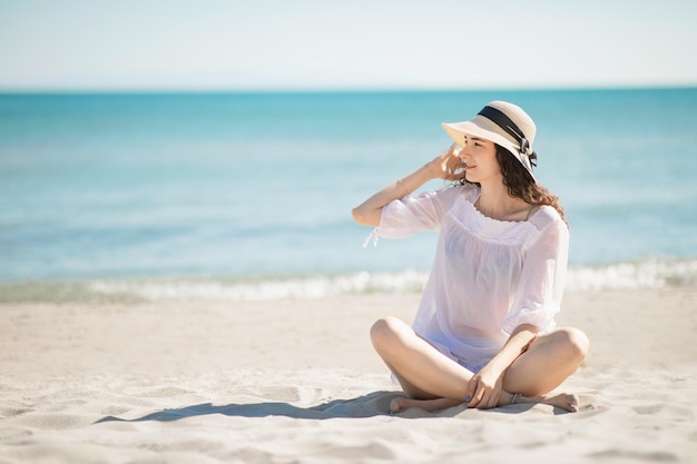 Belle adolescente adolescente assise sur la plage portant des vêtements de plage blancs et un chapeau de paille vacances d'été et concept de vacances