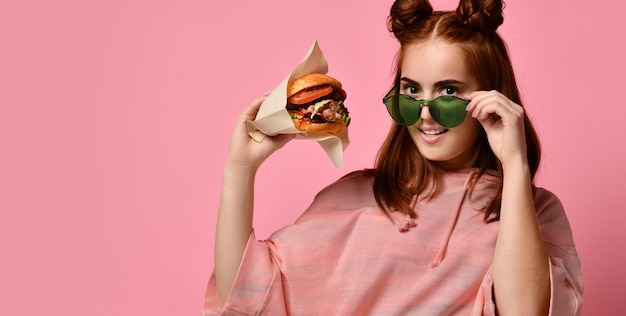 Belle adolescente aux cheveux rouges tenant un hamburger à la main une étudiante au gingembre a un déjeuner de restauration rapide