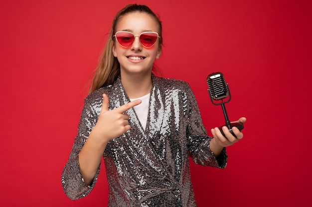 Belle adolescente brune positive portant une veste élégante et des lunettes rouges isolées