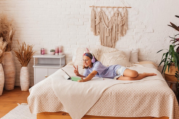 Belle adolescente communique en ligne par téléphone en position allongée sur le lit à la maison. L'enfant dans les écouteurs apprend en ligne en étant allongé sur le lit. Concept d'apprentissage à distance