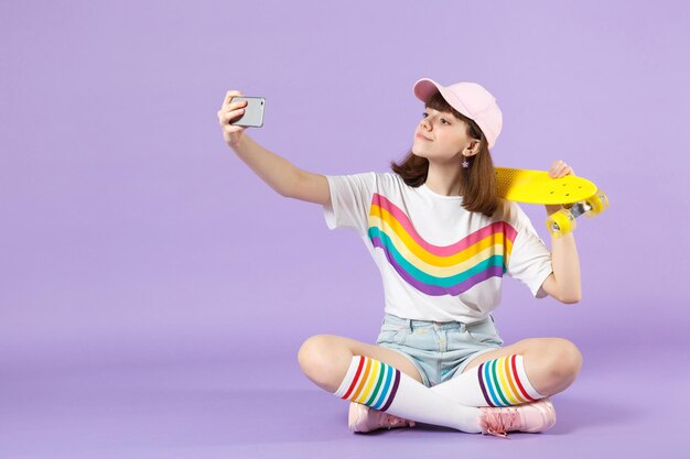 Belle adolescente dans des vêtements vifs tenant une planche à roulettes jaune, faisant un selfie tourné sur un téléphone portable isolé sur fond pastel violet. Émotions sincères des gens, concept de style de vie. Maquette de l'espace de copie.