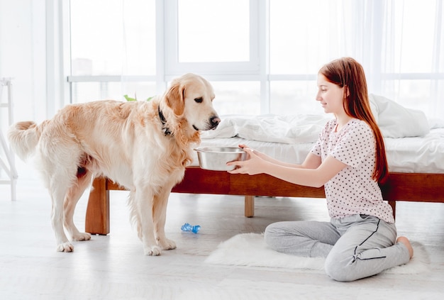 Belle adolescente donne de la nourriture au chien golden retriever dans la chambre le matin. Enfant nourrissant un chien dans la pièce avec la lumière du soleil. Relations amicales entre l'homme et l'animal