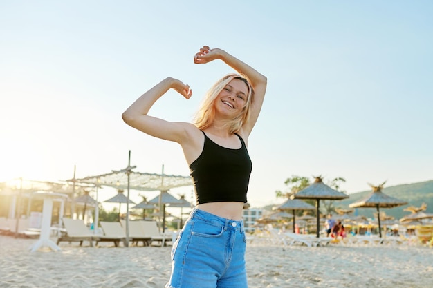 Belle adolescente heureuse sur une plage de sable avec les mains levées dansant au coucher du soleil