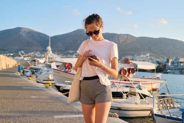 Belle adolescente marchant le long de la jetée avec des yachts amarrés, adolescent avec smartphone, fond de paysage de montagne coucher de soleil sur la mer, vacances d'été, voyage en mer