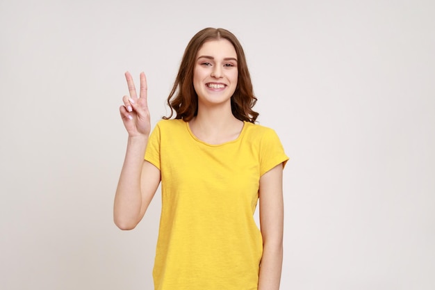 Belle adolescente satisfaite montrant le signe v symbole de paix avec les doigts, regardant la caméra avec un sourire heureux, célébrant le triomphe. Tourné en studio intérieur isolé sur fond gris.