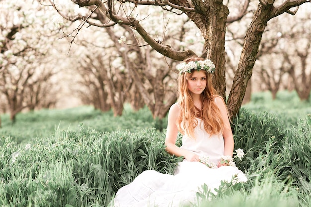 Belle adolescente se reposant dans un verger portant une robe élégante et une couronne de fleurs à l'extérieur