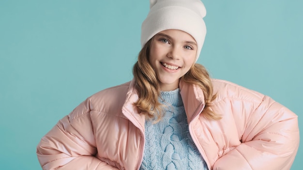 Belle adolescente vêtue d'une doudoune rose et d'un chapeau souriant sur fond bleu