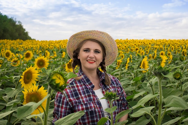 Belle agricultrice joyeuse et souriante d'âge moyen dans un chapeau de paille et une chemise se dresse dans un champ de récolte de tournesols par une journée ensoleillée