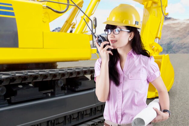 Une belle architecte parle avec un walkietalkie à côté d'un tracteur sur un chantier de construction