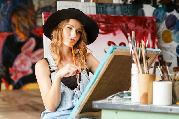 Belle artiste féminine aux cheveux longs dans un élégant chapeau noir tenant une toile et un pinceau et regardant la peinture
