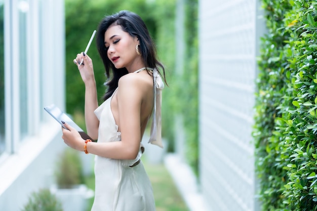 Belle asiatique freelance Success people femme d'affaires mannequin en robe d'été blanche sans manches utilisant un travail décontracté avec une tablette pour naviguer sur Internet, discuter et bloguer dans un café