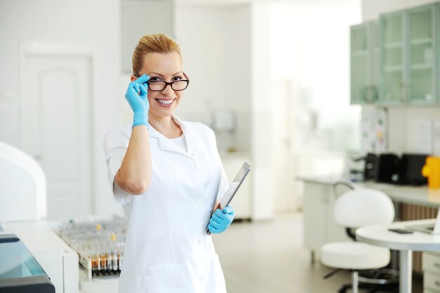 Belle assistante de laboratoire blonde en blouse blanche et gants en caoutchouc debout en laboratoire avec tablette dans ses bras.