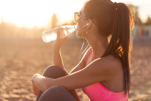 Belle athlète de remise en forme femme buvant de l'eau après l'entraînement s'exerçant le soir du coucher du soleil l'été dans le portrait en plein air de la plage