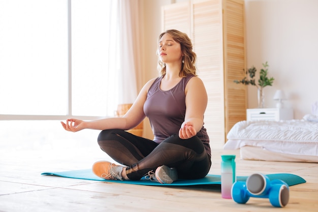 Belle belle femme assise dans la pose de lotus tout en pratiquant le yoga à la maison