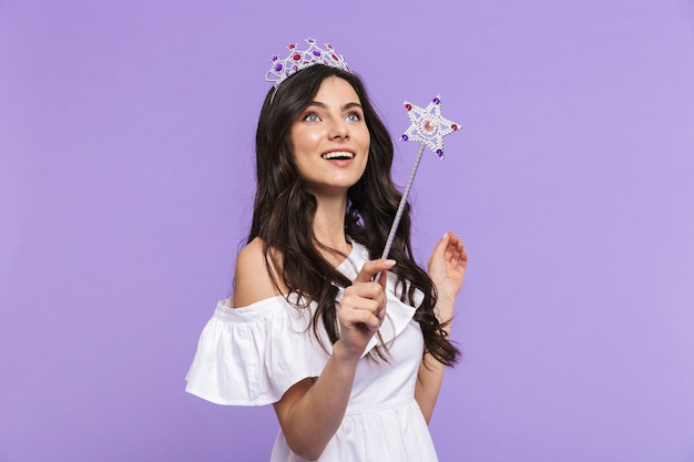 Belle belle jeune femme portant une tenue de princesse isolée sur un mur violet, posant avec une baguette magique