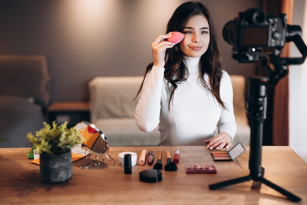 Belle blogueuse beauté filmant un tutoriel de routine de maquillage quotidien à la caméra. Influenceuse jeune femme en direct, revue de produits cosmétiques en streaming dans un home studio. Travail de blogueur. DIY se maquiller.