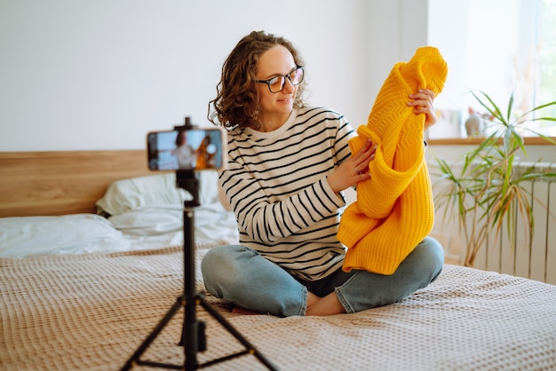 Belle blogueuse de mode tenant un pull jaune enregistrant rêveusement une nouvelle vidéo pour vlog en ligne