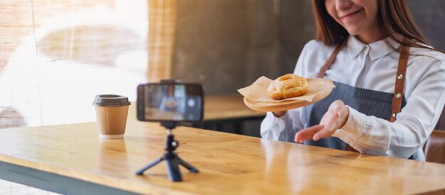 Une belle blogueuse ou vlogger culinaire asiatique montrant un morceau de beignet tout en enregistrant une vidéo devant une caméra