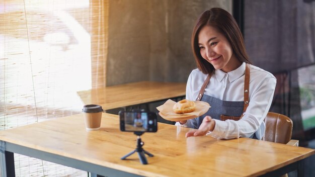 Une belle blogueuse ou vlogger culinaire asiatique montrant un morceau de beignet tout en enregistrant une vidéo devant une caméra