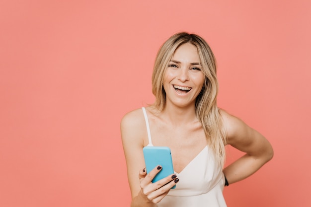 Belle blonde en blouse blanche en riant après avoir lu des histoires drôles sur smartphone. Les choses rendent heureux. Sur fond rose avec un espace de copie. Concept d'émotions de personnes.