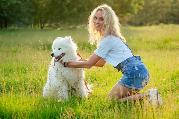 Belle blonde bouclée souriante heureuse jeune femme en short en jean s'entraînant et jouant un chien samoyède mignon et moelleux blanc dans le parc d'été fond de champ de rayons de coucher de soleil. animal de compagnie et hôtesse.