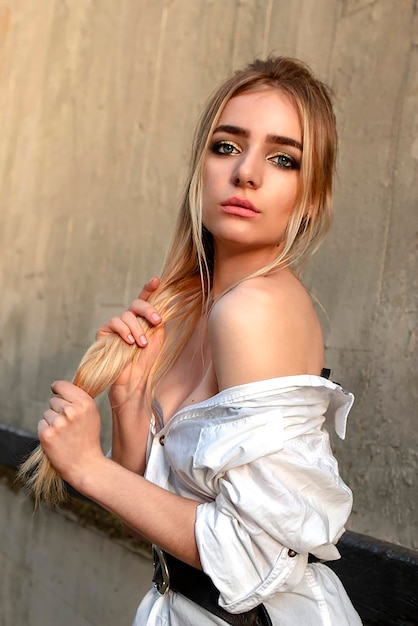 Belle blonde en chemise blanche tient les cheveux dans ses mains. posant sur un fond de mur