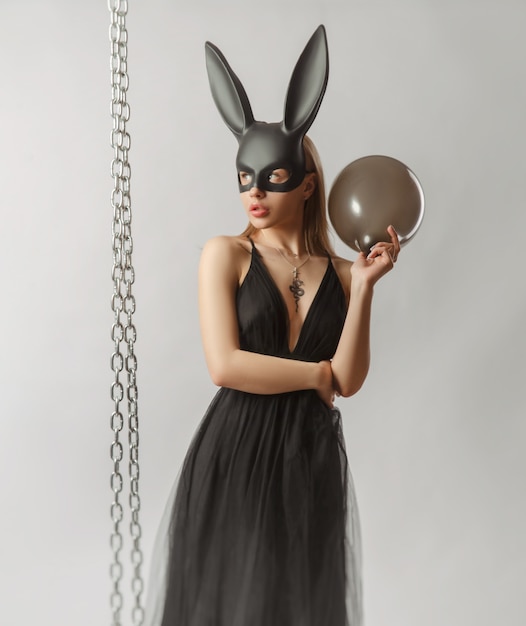 La belle blonde dans un masque de lapin dans une robe de soirée sexy sur fond blanc avec une chaîne en métal