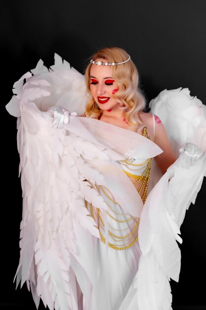 Belle blonde dans une robe blanche sexy ornée de perles dorées et de grandes ailes blanches dans le dos Traces de baisers sur le corps et le visage Cupidon sexy Fond noir