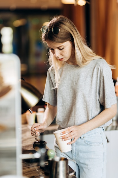Une belle blonde mince aux cheveux longs, vêtue d'une tenue décontractée, prépare du café dans un café moderne. Le processus de préparation du café est montré. .