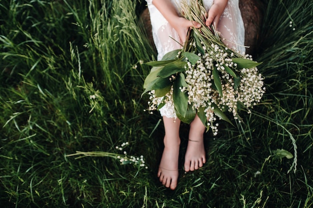 Une belle blonde de neuf ans dans une longue robe blanche, tenant un bouquet de muguets, gros plan d'une jeune fille tenant des fleurs de la vallée.Été, coucher de soleil.