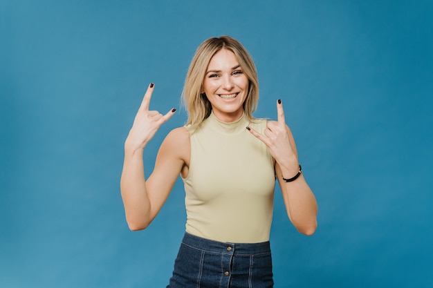 Belle blonde positive dans un t-shirt léger et une jupe en jean, montre un large geste de main rock souriant, sur fond bleu avec copie espace. Émotions sincères.