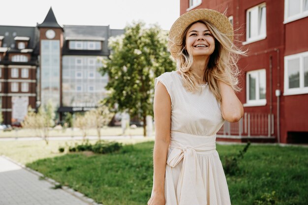 Une belle blonde se promène dans une ville européenne. Femme en robe blanche et chapeau de paille, elle sourit et est heureuse