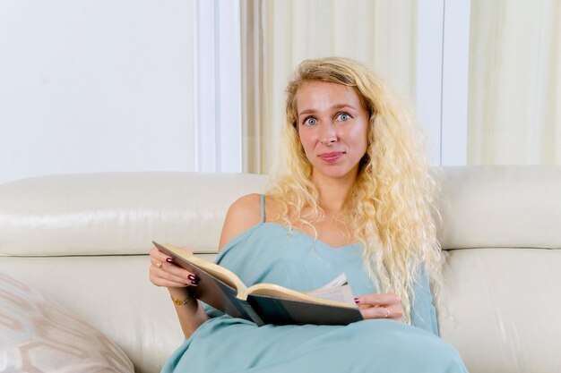 Belle blonde souriante se détendre sur le canapé et lire un livre