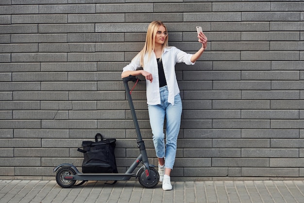 Belle blonde en vêtements décontractés debout avec scooter électrique à l'extérieur pendant la journée ensoleillée avec téléphone dans les mains