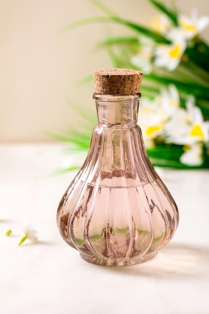 Belle bouteille transparente rose de parfum avec du liège debout sur une table en marbre blanc Petites fleurs blanches jonquilles de printemps à l'arrière-plan Concept de fabrication de parfum avec place pour l'étiquette de conception