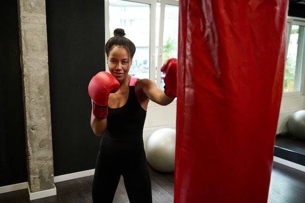 Belle boxeuse africaine portant des gants de boxe rouges frappe sur un sac de boxe, regarde la caméra tout en effectuant des arts martiaux de combat dans une salle de sport