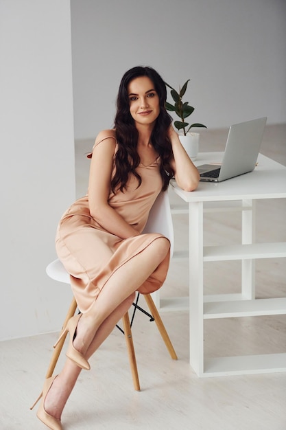 Belle brune aux cheveux bouclés dans des vêtements de luxe est assise à table avec un ordinateur portable à la maison pendant la journée