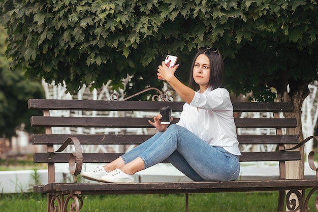 belle brune sur un banc de parc avec des verres de café et prend un selfie au téléphone