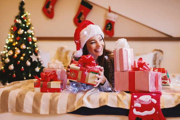 Belle brune avec un chapeau de père Noël sur la tête posant dans son lit entouré de cadeaux de Noël.