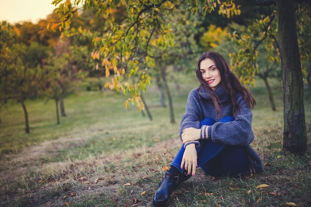 Belle brune dans un pull en tricot assis sous l'arbre dans un parc d'automne