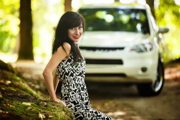 Belle brune dans une robe posant dans la nature près d'une voiture blanche.