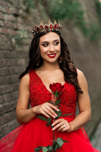 Belle brune dans une robe rouge avec une couronne. Fille brune avec une rose rouge à l'extérieur sur mur loft en pierre