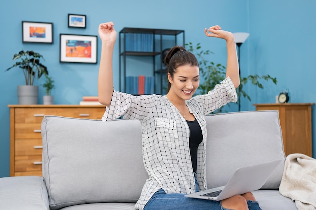 Une belle brune est assise sur un canapé à la maison avec un ordinateur portable sur les genoux en train de discuter avec une webcam