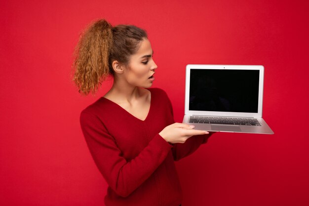 Belle charmante fascinante étonnée jeune femme blonde foncée bouclée portant un pull rouge debout